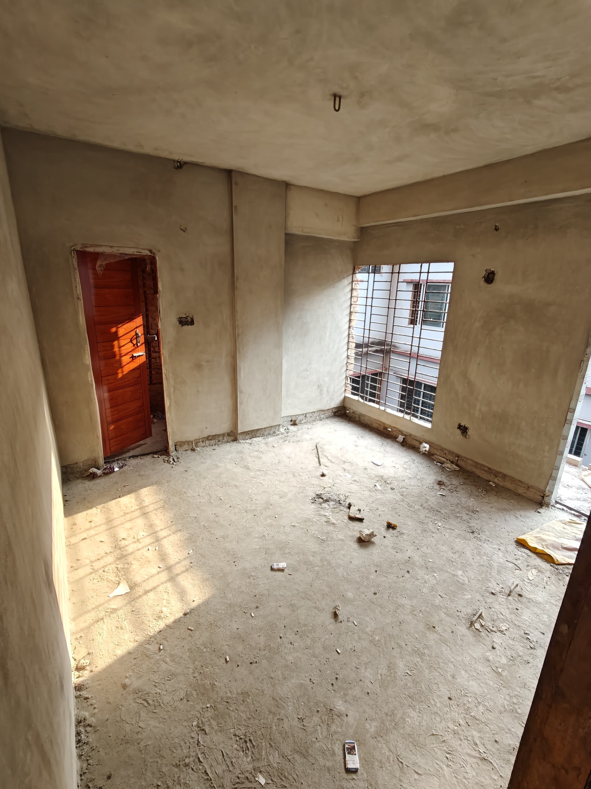 Sunlight streams into an unfinished concrete room with a wooden door and barred window.