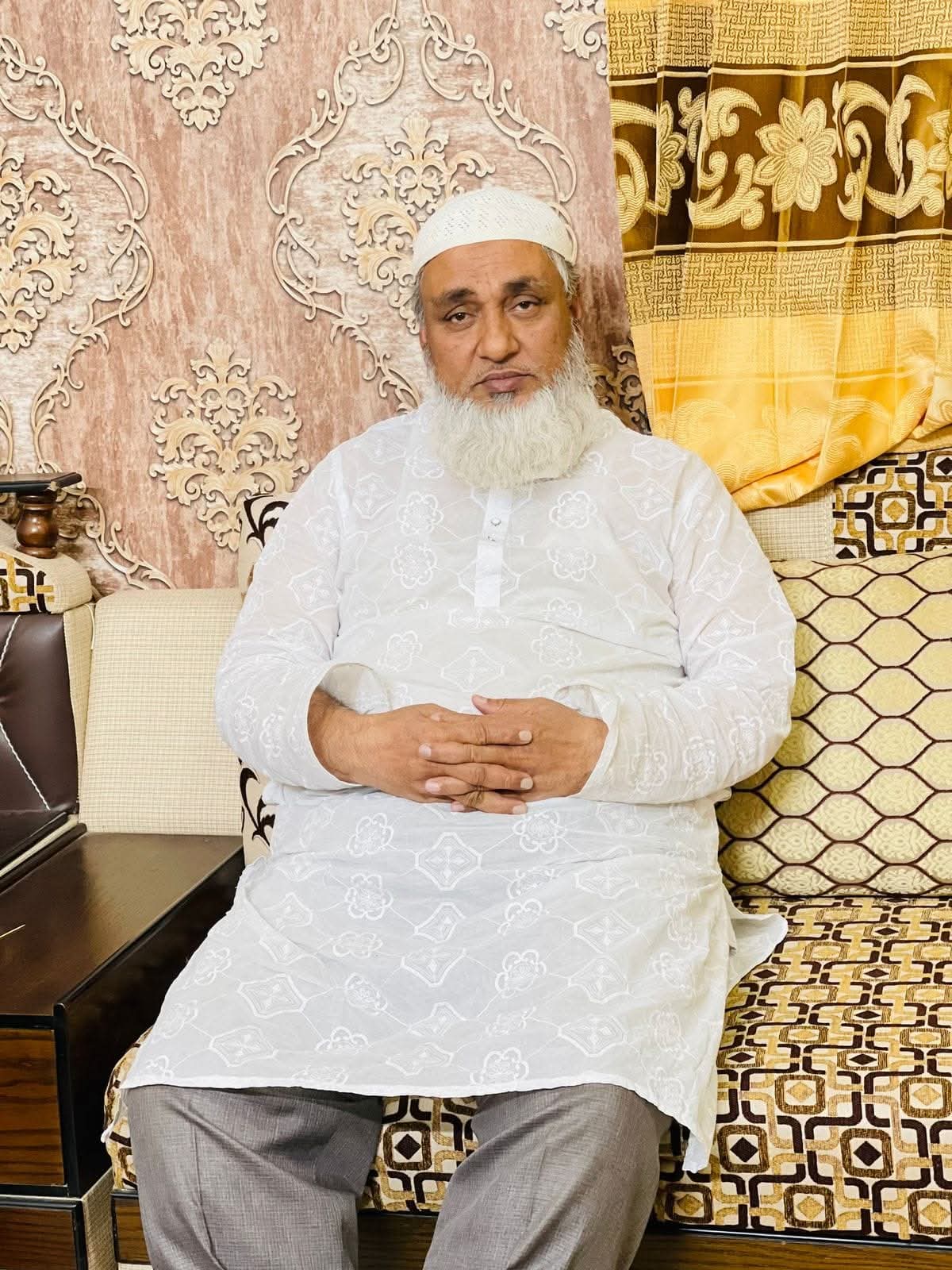 Elderly man with a white beard and skullcap sitting on an ornate patterned sofa.