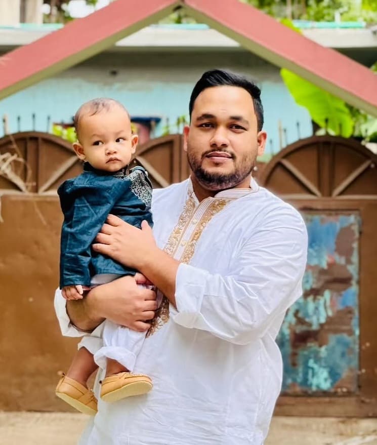 Bearded man in a white embroidered tunic holding a baby boy in a blue outfit.